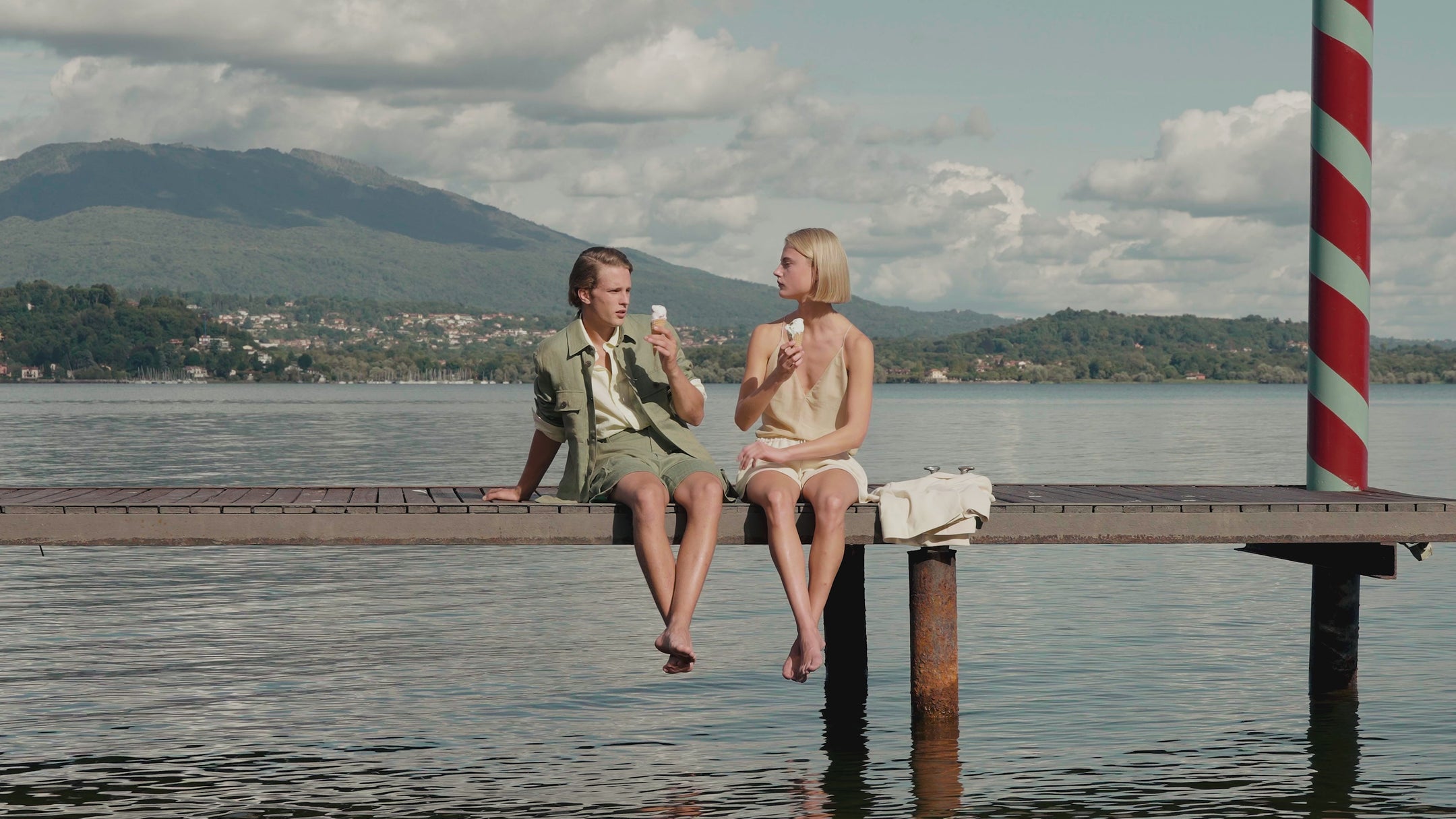 Two young adults wearing casual summer outfits while enjoying ice cream on a lakeside pier.