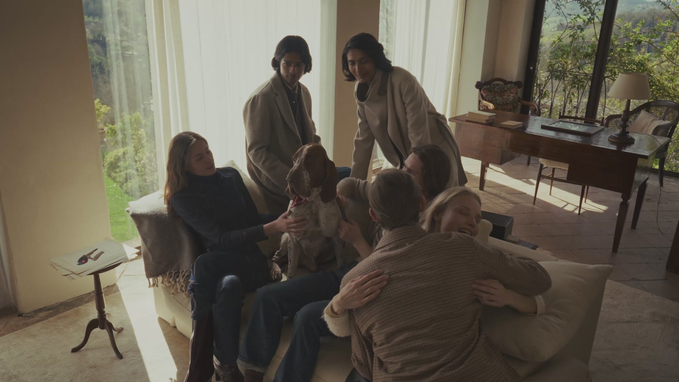 Group of people gathered in a sunlit living room petting a dog on a sofa, wearing casual and tailored coats in neutral tones