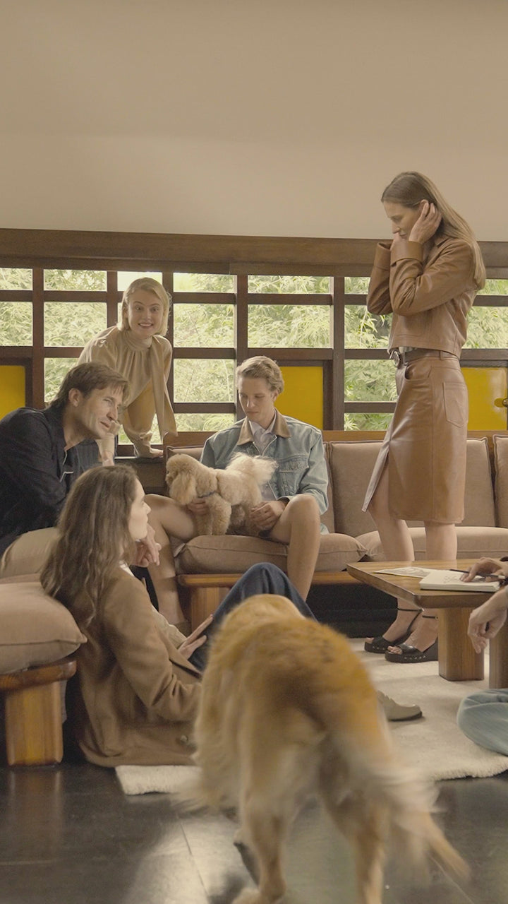A group of friends gathered in a mid-century modern living room, relaxed and laughing together, with two dogs roaming freely around them