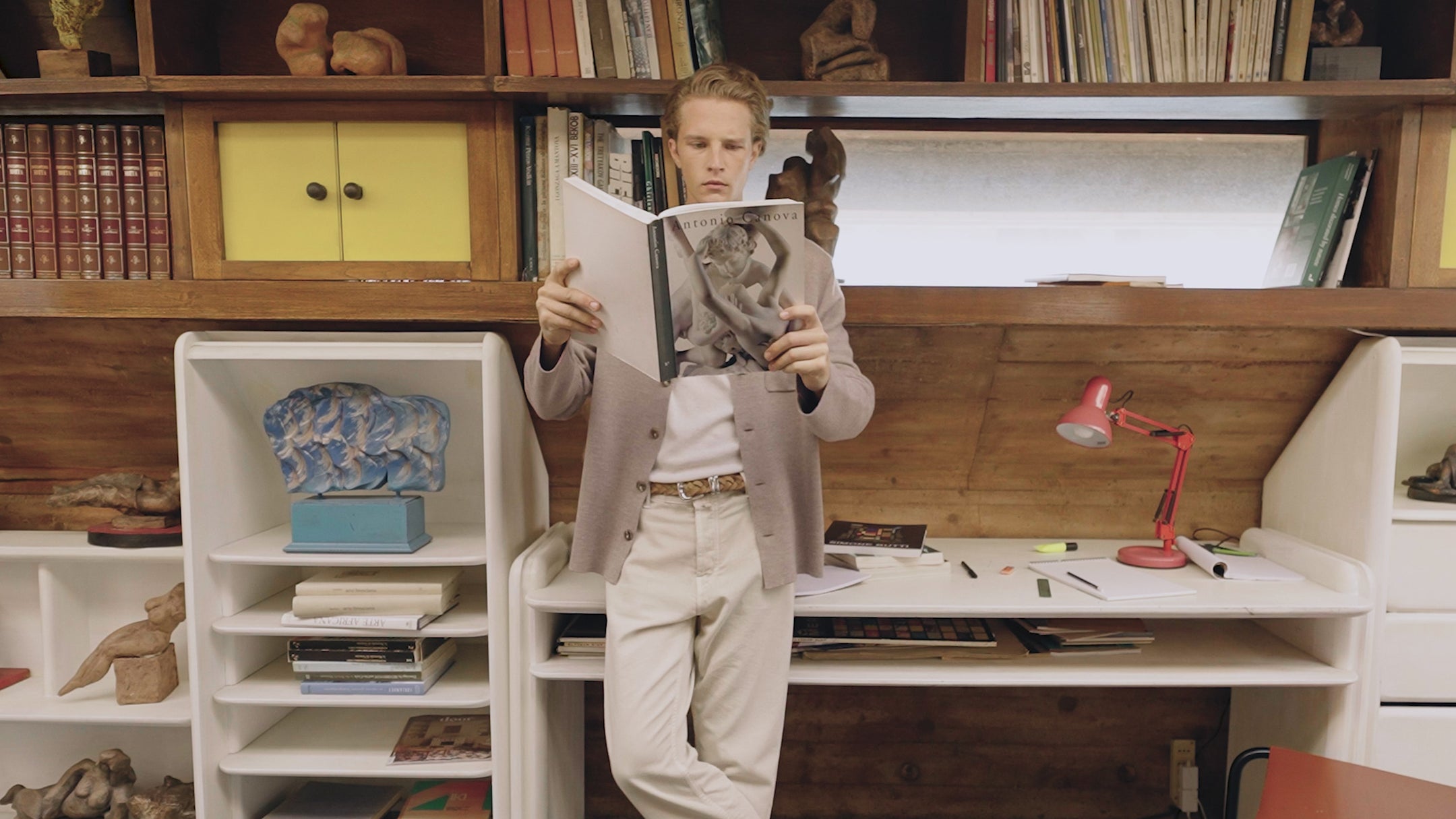 Individual wearing Jacob Cohën clothing while standing in a study-like interior, reading a large book in front of a wall of shelves filled with books and decorative objects