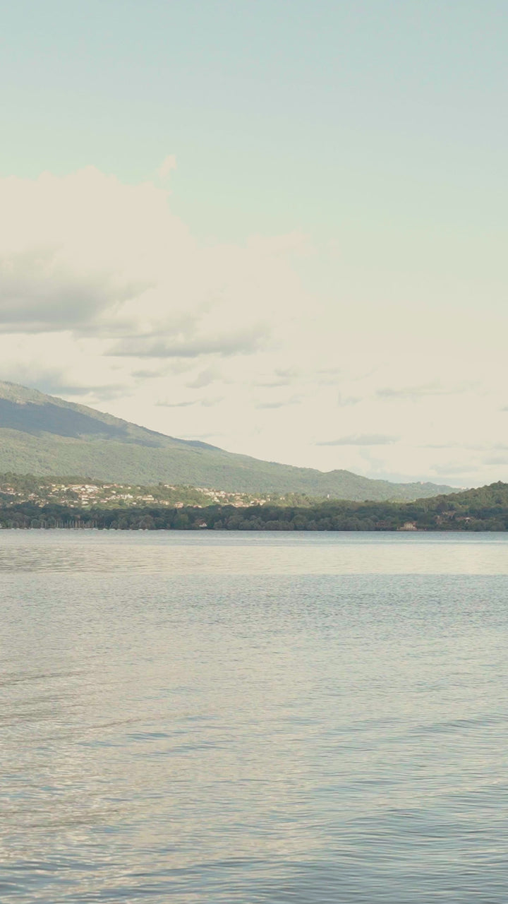 Two people standing on a lakeside dock, viewed from behind, wearing linen shirts in light purple and aqua blue with striped bermuda swimwear, holding wooden polo mallets, with a serene mountain lake landscape in the background.