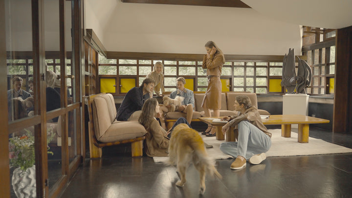 Fashion campaign video featuring a group of models gathered in a mid-century modern living room, wearing a mix of tailored and casual looks in neutral tones — including a camel coat, denim jacket, and dark separates — interacting naturally with each other and a golden retriever dog
