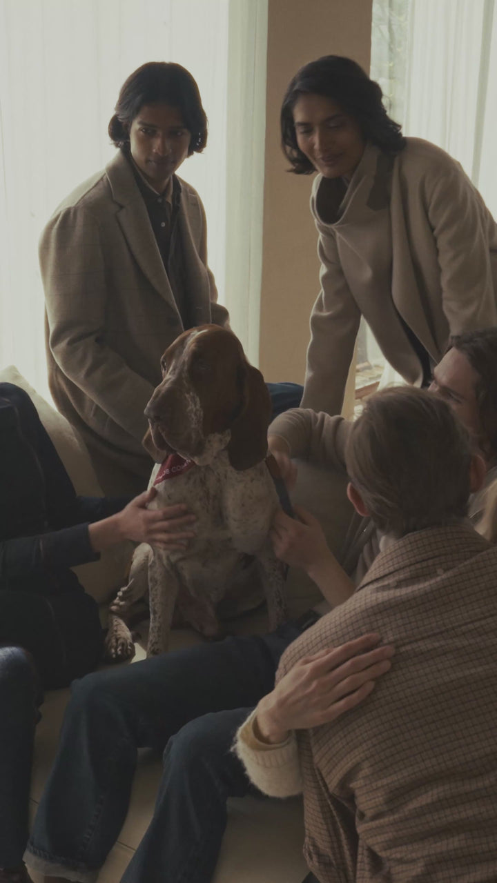 Group of people gathered in a sunlit living room petting a dog on a sofa, wearing casual and tailored coats in neutral tones