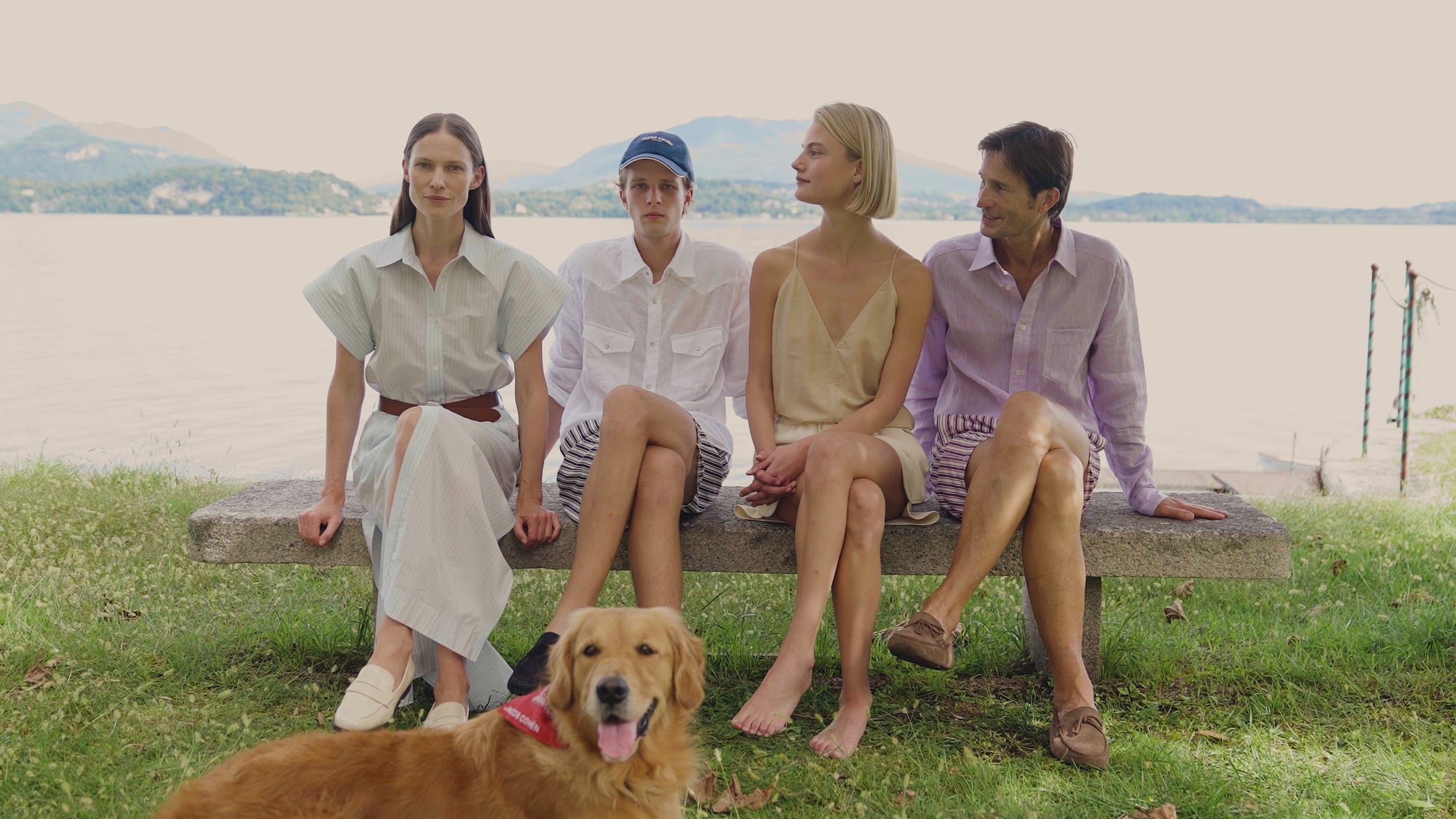 Four models sitting on a stone bench by a lake, wearing summer outfits in neutral and pastel tones — linen shirts, swim shorts and a slip dress. A golden retriever lies on the grass in the foreground. Mountain landscape in the background