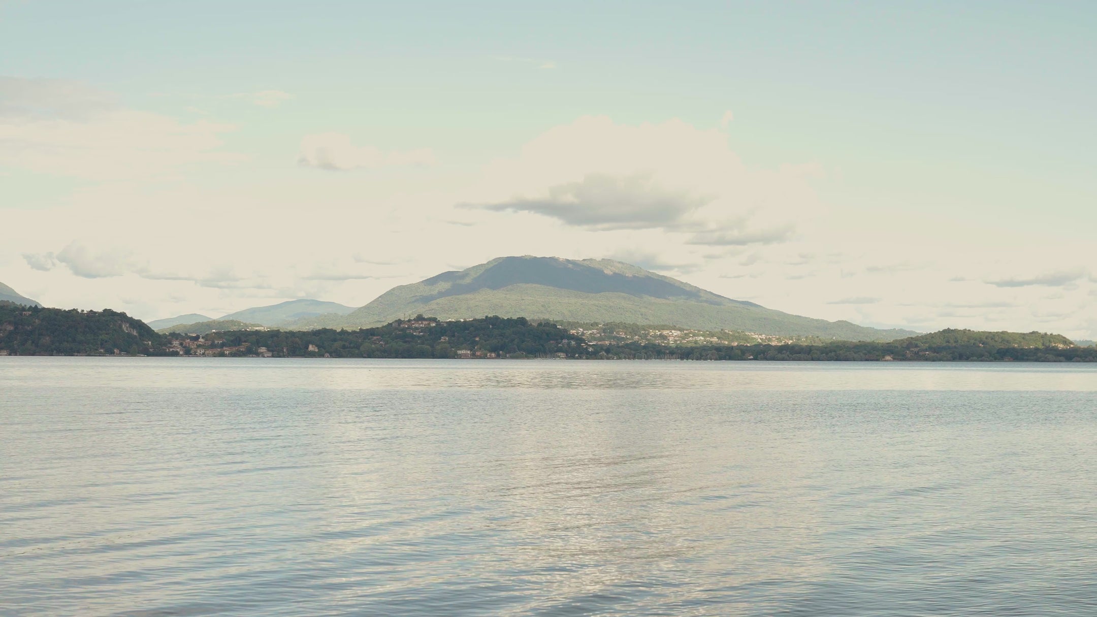 Two people standing on a lakeside dock, viewed from behind, wearing linen shirts in light purple and aqua blue with striped bermuda swimwear holding wooden polo mallets, with a serene mountain lake landscape in the background.
