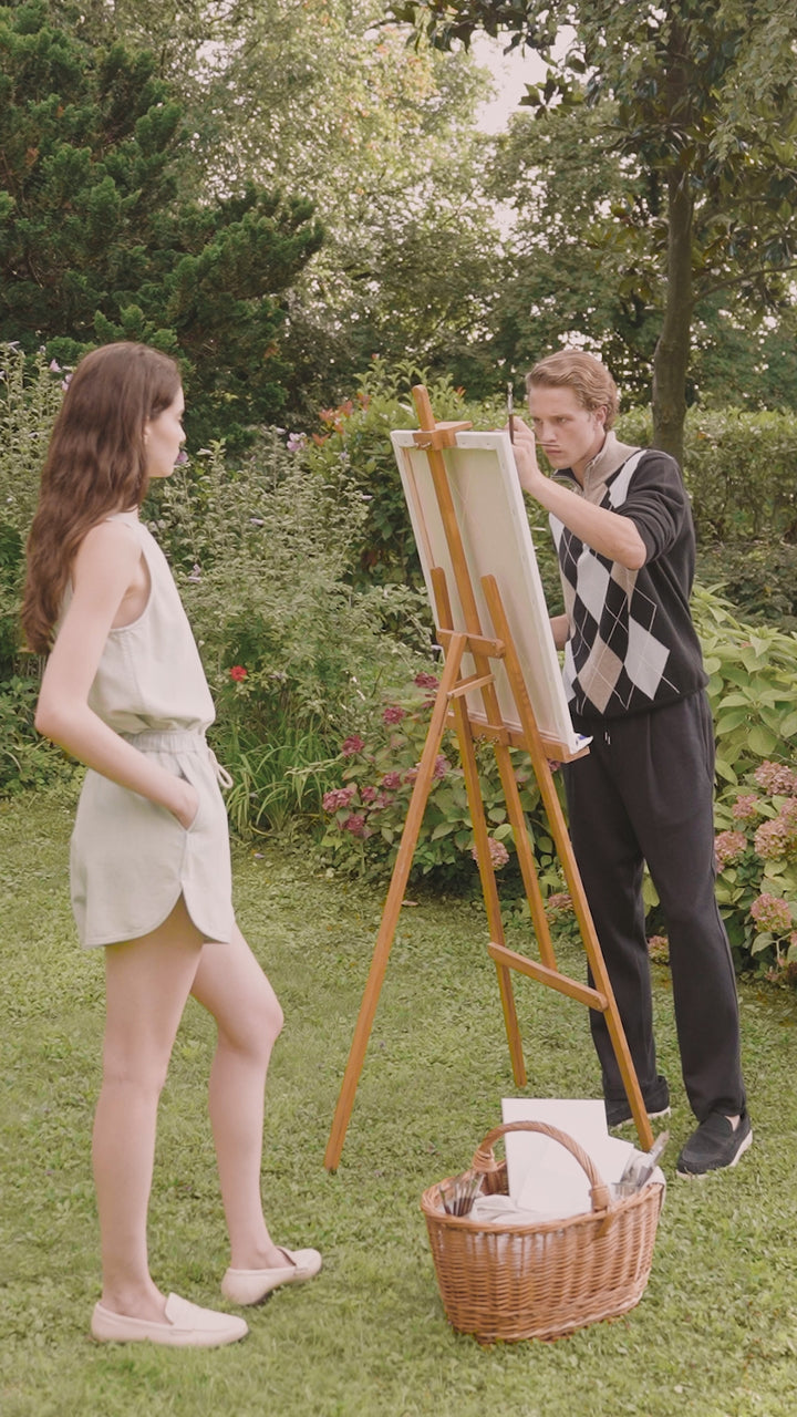 Two people are painting together outdoors in a beautiful garden. She's wearing a light blue outfit with shorts and a tank top, he's in a sweater and chino pants. They're surrounded by colorful flower beds with orange-red blooms, green lawns, and trees. A picnic basket sits at their feet. A romantic, relaxing scene in nature