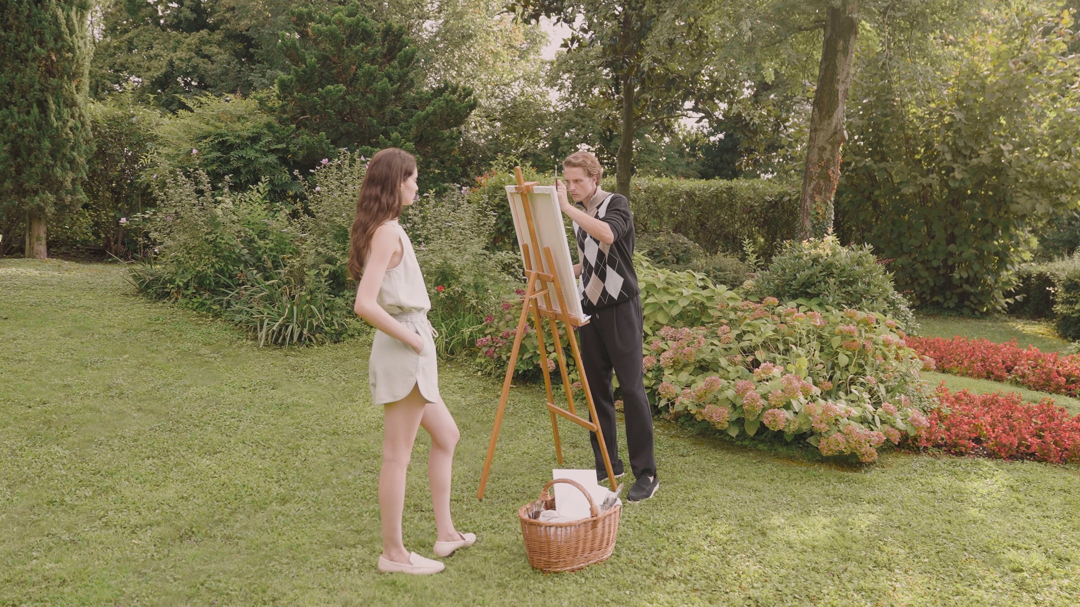 Two people are painting together outdoors in a beautiful garden. She's wearing a light blue outfit with shorts and a tank top, he's in a sweater and chino pants. They're surrounded by colorful flower beds with orange-red blooms, green lawns, and trees. A picnic basket sits at their feet. A romantic, relaxing scene in nature